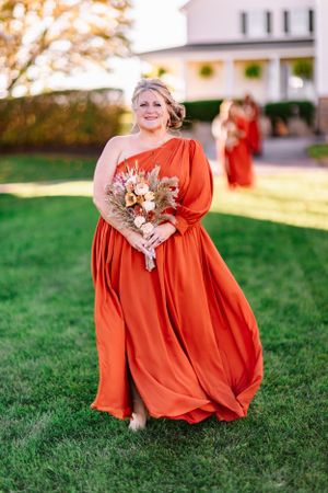 Bridesmaid in one-shoulder orange gown holds bouquet while standing on grass with white farmhouse behind.