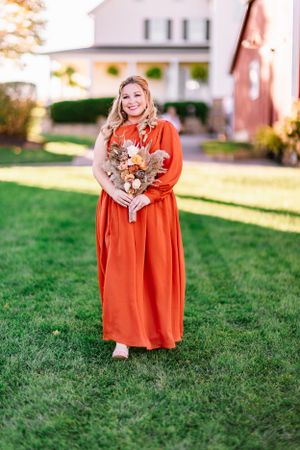 Bridesmaid poses with dried flower bouquet in orange dress on lawn with farmhouse in background.