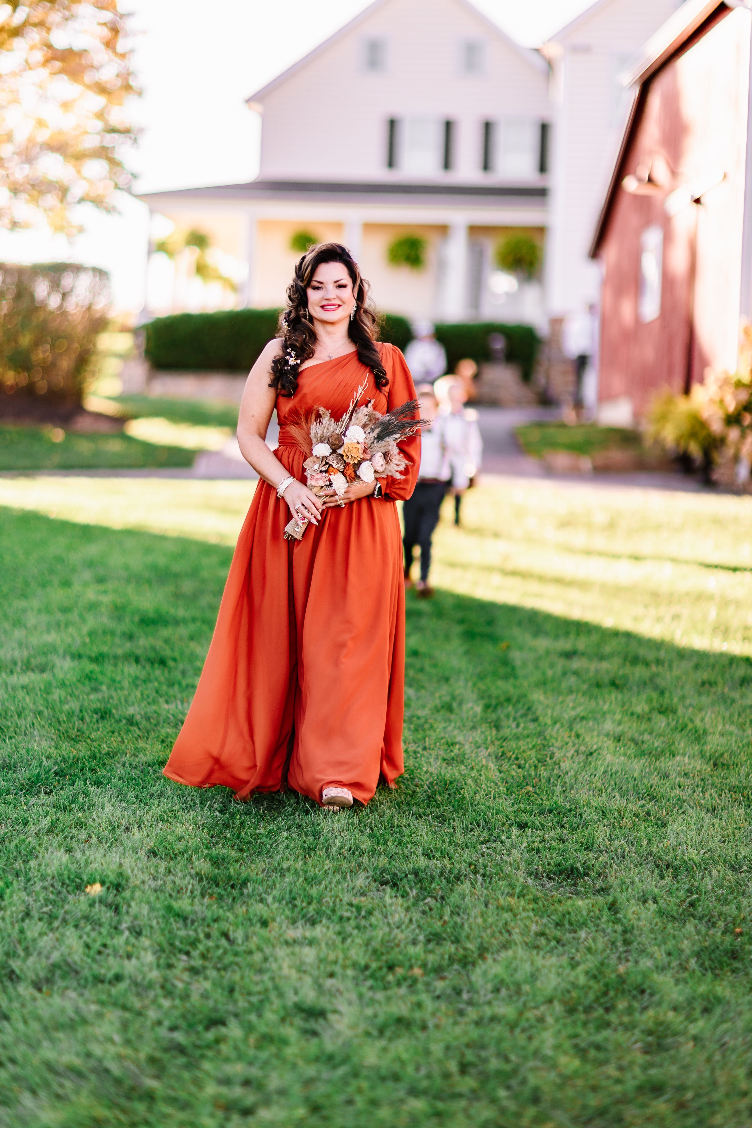 Woman in flowing burnt orange dress holds a bouquet against white farmhouse backdrop.