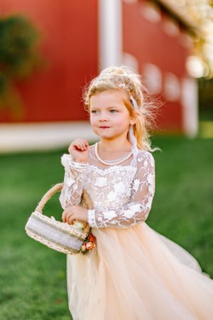 A young flower girl in a delicate white lace and tulle dress carries a small basket outdoors.