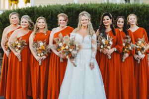 Bridal party in matching orange dresses poses with bride in lace gown holding fall bouquets.