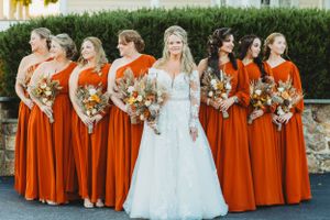 Bride poses with bridesmaids wearing vibrant orange dresses holding fall-themed bouquets.