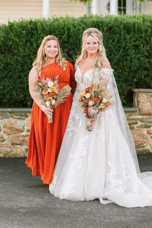 Two women with fall-themed bouquets pose together at outdoor wedding venue.