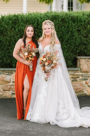 Wedding photo of two women with bouquets against stone wall and greenery backdrop.