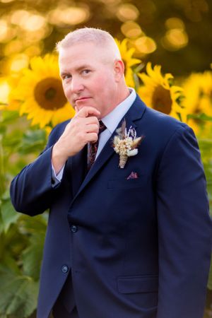 Groom in classic navy suit poses thoughtfully against sunflower garden backdrop.