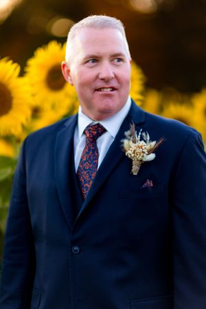 Groom wearing navy suit and patterned tie stands in front of blooming sunflower field.