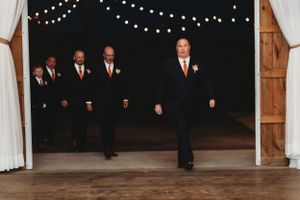 Wedding party members in formal attire walk through rustic barn entrance with string lights.