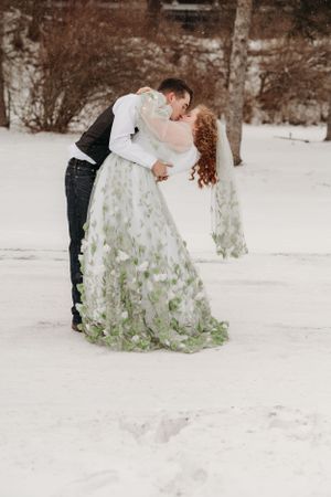 Winter wedding scene with couple in white dress with green details against snowy background.