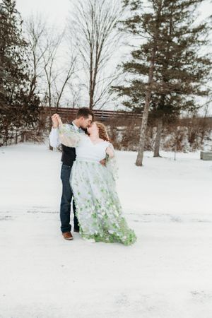 Couple shares romantic moment in snowy winter setting wearing white and green floral dress.