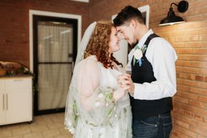 Romantic indoor dance moment with couple in white dress and vest against brick wall.