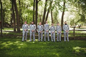 Wedding party in white and light gray suits posing in a line on grass with trees in background.