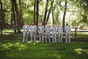 Wedding party wearing light colored suits stand in a row outdoors among trees.