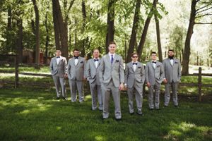 Groomsmen in gray suits arranged in a V formation in a sunlit wooded area.