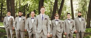 A wedding party of groomsmen in light gray suits pose together in a wooded setting.