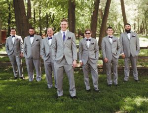 Wedding party in coordinated gray suits standing in V-formation on grass with trees behind.