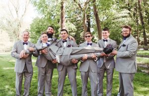Groomsmen in gray suits playfully holding up one member of the wedding party for a group photo.