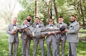 A groomsmen group in matching gray suits strike a playful pose while holding up their friend in an outdoor setting.