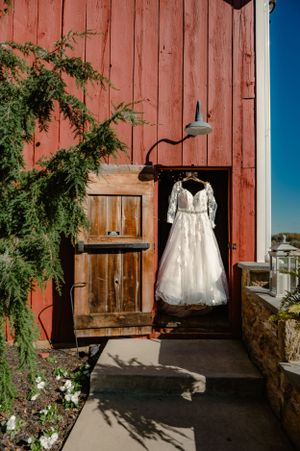 A white wedding dress hangs elegantly against a red barn exterior with rustic wooden doors.