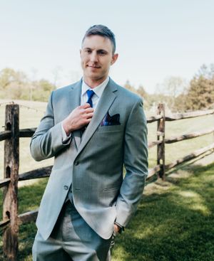 A formal portrait of a man in a gray suit and blue tie standing by a rustic wooden fence.