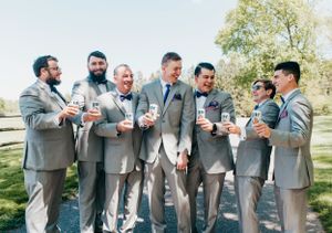 Groomsmen sharing laughs and drinks while celebrating outdoors in matching gray suits.