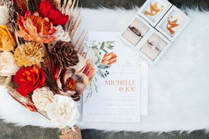 Wedding invitation with floral arrangement featuring red and peach flowers on white fur background.