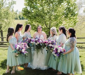 Wedding party gathered outdoors holding pink and purple floral bouquets under green trees.