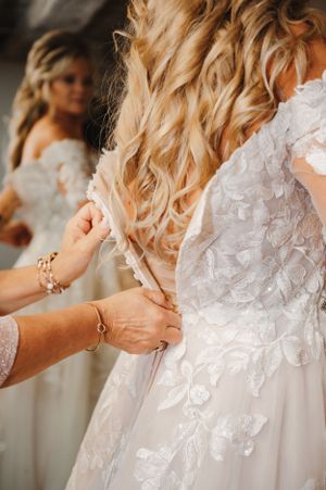 Delicate lace details and buttons being fastened on the back of an ornate white wedding gown.