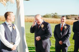 Emotional wedding ceremony moment under white arch with groomsmen standing by.