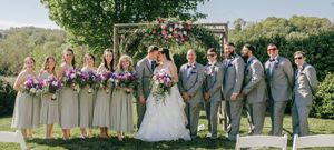 Full wedding party portrait with bridesmaids in sage and groomsmen in gray standing on lawn.