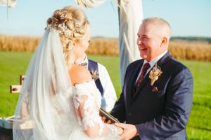 Heartfelt exchange during outdoor wedding ceremony against rural backdrop.