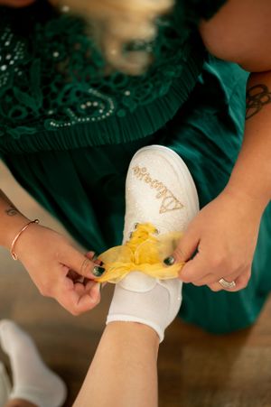 Hands tying yellow ribbon on white bridal shoe with emerald green dress in background.