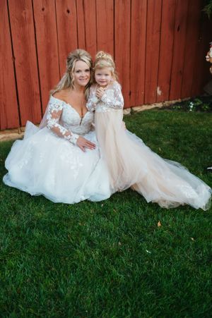 Bride in lace gown poses with flower girl in matching dress against red barn.