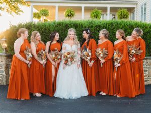 A bridal party wearing bright orange dresses poses with the bride in white against a porch backdrop.