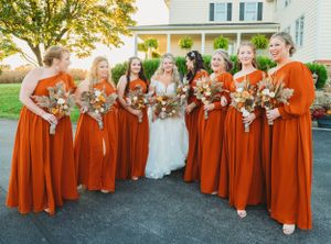 Bride and bridesmaids in burnt orange dresses pose together outside white house with fall bouquets.