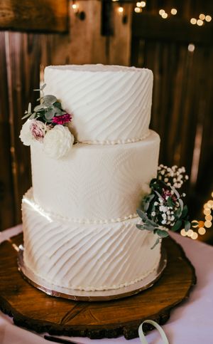 Elegant three-tier white wedding cake with diagonal texture and floral accents on a wooden stand.