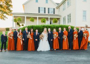 Full wedding party in orange and navy attire lined up for formal photo outside white colonial home.