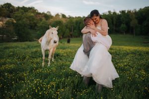 A romantic wedding photo with a white horse grazing in a flower-filled meadow at sunset.