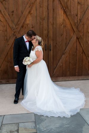 Romantic wedding kiss photographed against rustic barn doors with flowing white dress train.