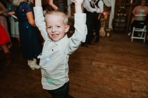 Young child in light colored shirt dances with arms raised at wedding reception.