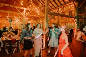 Wedding reception guests mingle and dance in a warmly lit rustic barn venue.