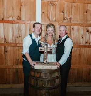 Wedding party poses with drinks around rustic wooden barrel in barn setting.