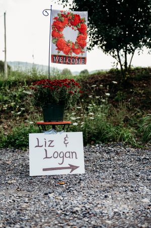 Wedding directional sign with red floral welcome flag on a gravel path.
