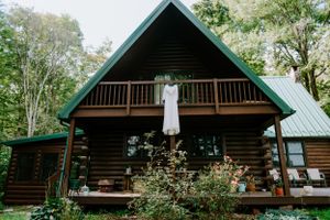 White wedding dress hangs on rustic log cabin balcony surrounded by lush green forest.