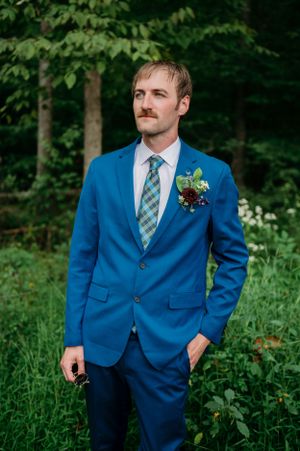 Groom in vibrant blue suit with plaid tie and boutonniere poses in wooded setting.