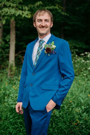 Groom smiling in bright blue suit with plaid tie against green forest background.