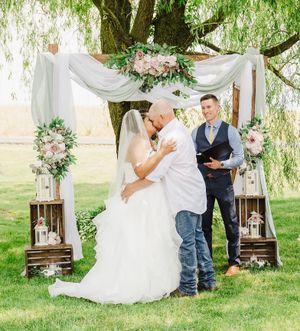 A couple embraces under a draped floral arch during their outdoor wedding ceremony.