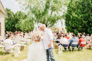 Outdoor wedding ceremony at a rustic venue with white folding chairs and guests seated under green trees.