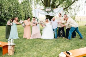 Wedding party strikes playful poses under a willow tree with white drapery and floral decorations.