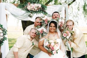 Wedding party posing joyfully under a white floral arch decorated with pink roses.