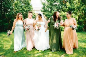 Bridesmaids in soft pastel dresses stand with bride on grassy lawn during summer wedding.